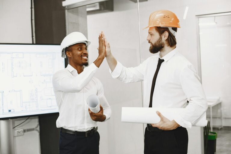 Two engineers in safety helmets celebrate a successful plan with a high-five indoors.