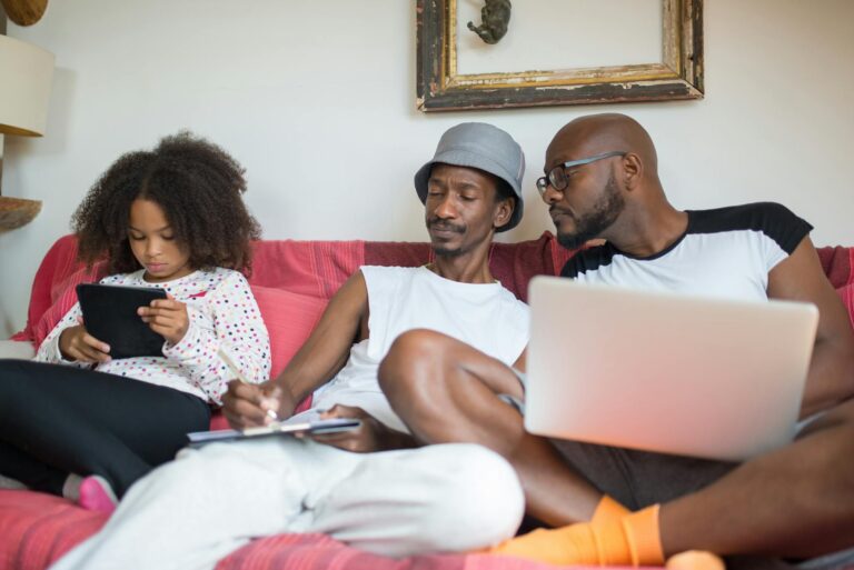 A family of three bonding on the couch with laptops and tablets in a cozy indoor setting.