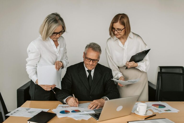 Three mature professionals collaborate at an office desk, analyzing charts and documents.