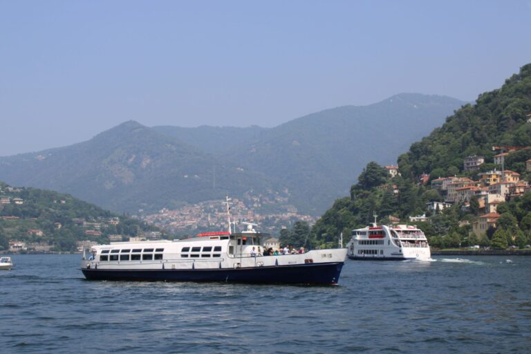 Passenger boats navigate the serene waters of Lake Como under the lush hills and clear blue sky.