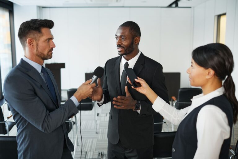 African American businessman in formal attire being interviewed by journalists with microphones in a conference room.